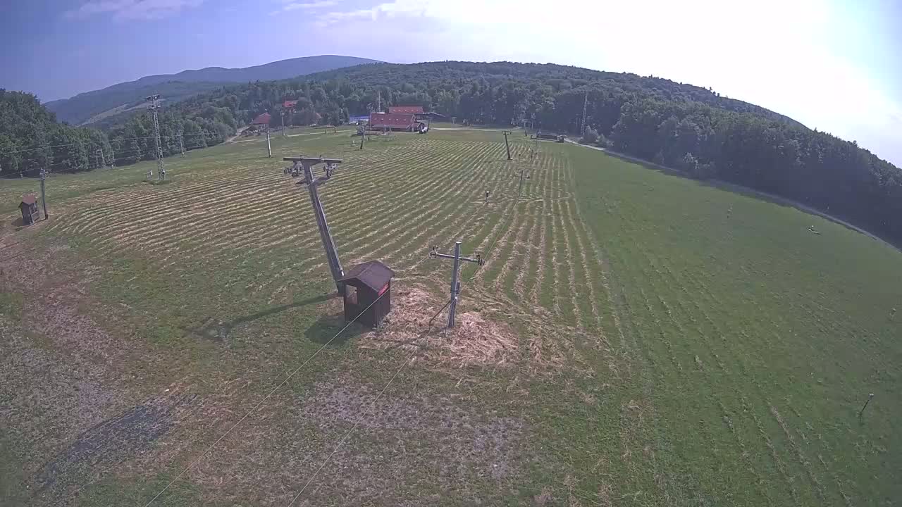 A grassy field with mowed lines, two utility poles, and a small wooden structure is visible, under a mostly sunny sky with distant mountains in the background.