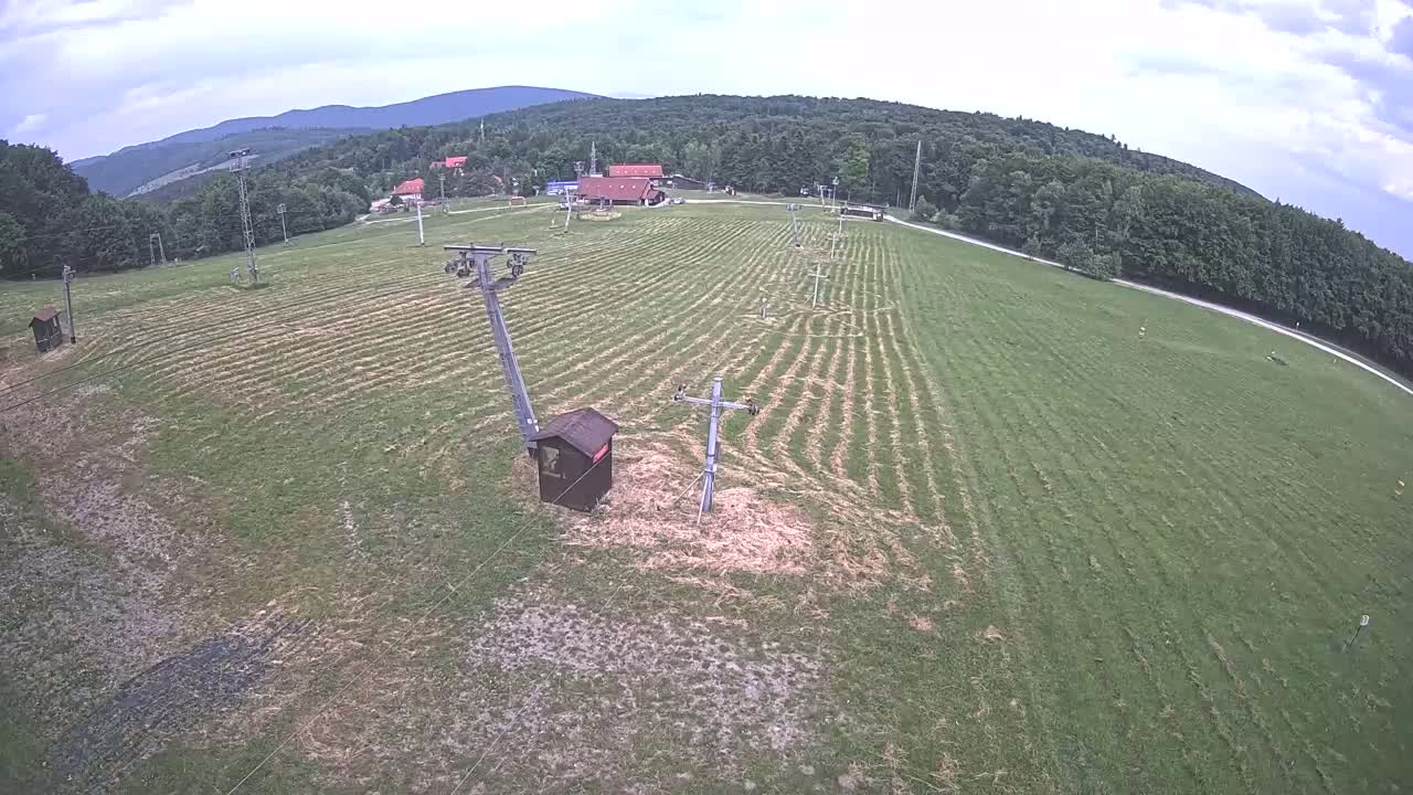 An aerial view shows a grassy field with parallel lines, two ski lift poles, a small building, and a line of trees and buildings in the background under a partly cloudy sky.