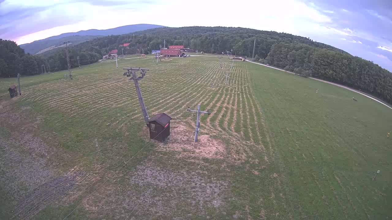 An aerial view shows a grassy field with parallel lines, two utility poles, a small wooden structure, and a forested hillside in the background under a partly cloudy sky.