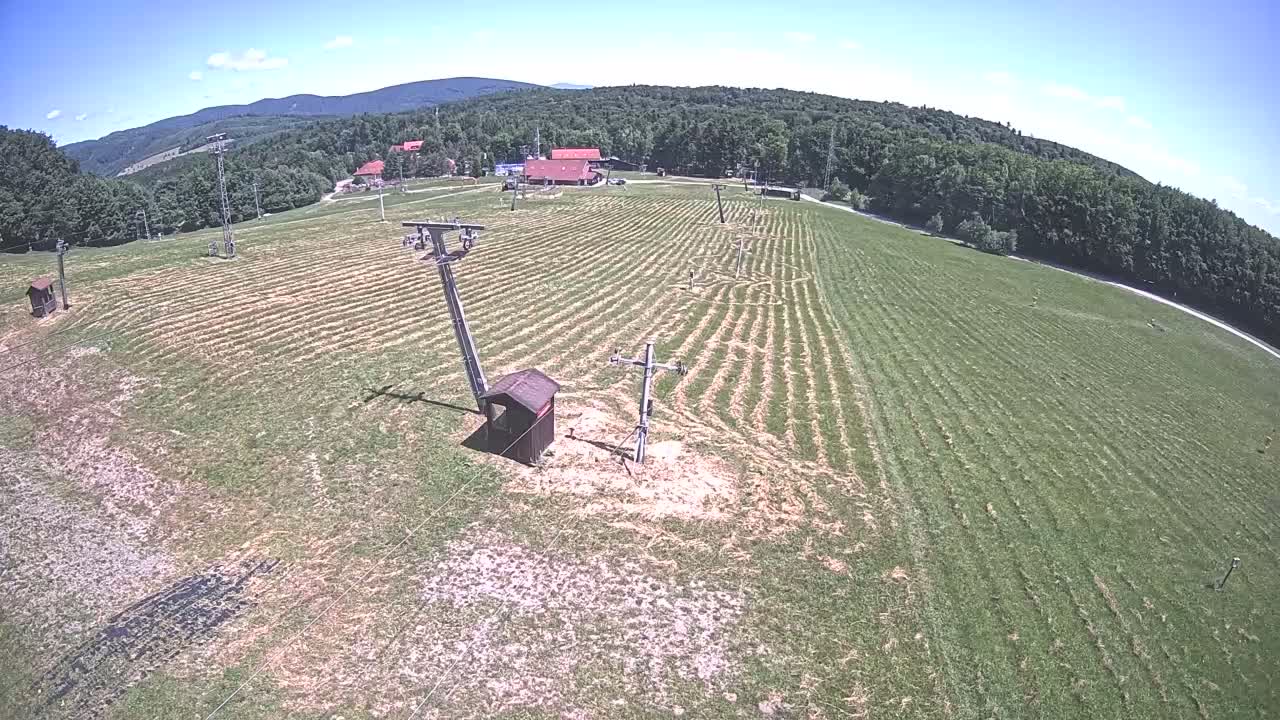 An aerial view shows a mown grassy field with ski lift infrastructure, a small building, and a background of forested hills under a sunny sky.