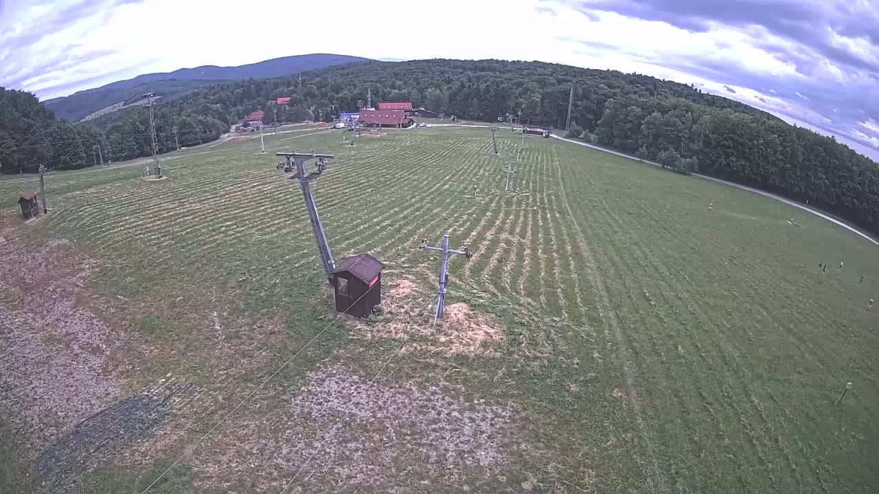 An aerial view shows a grassy field with a ski lift infrastructure, a few buildings in the distance, and a partly cloudy sky.