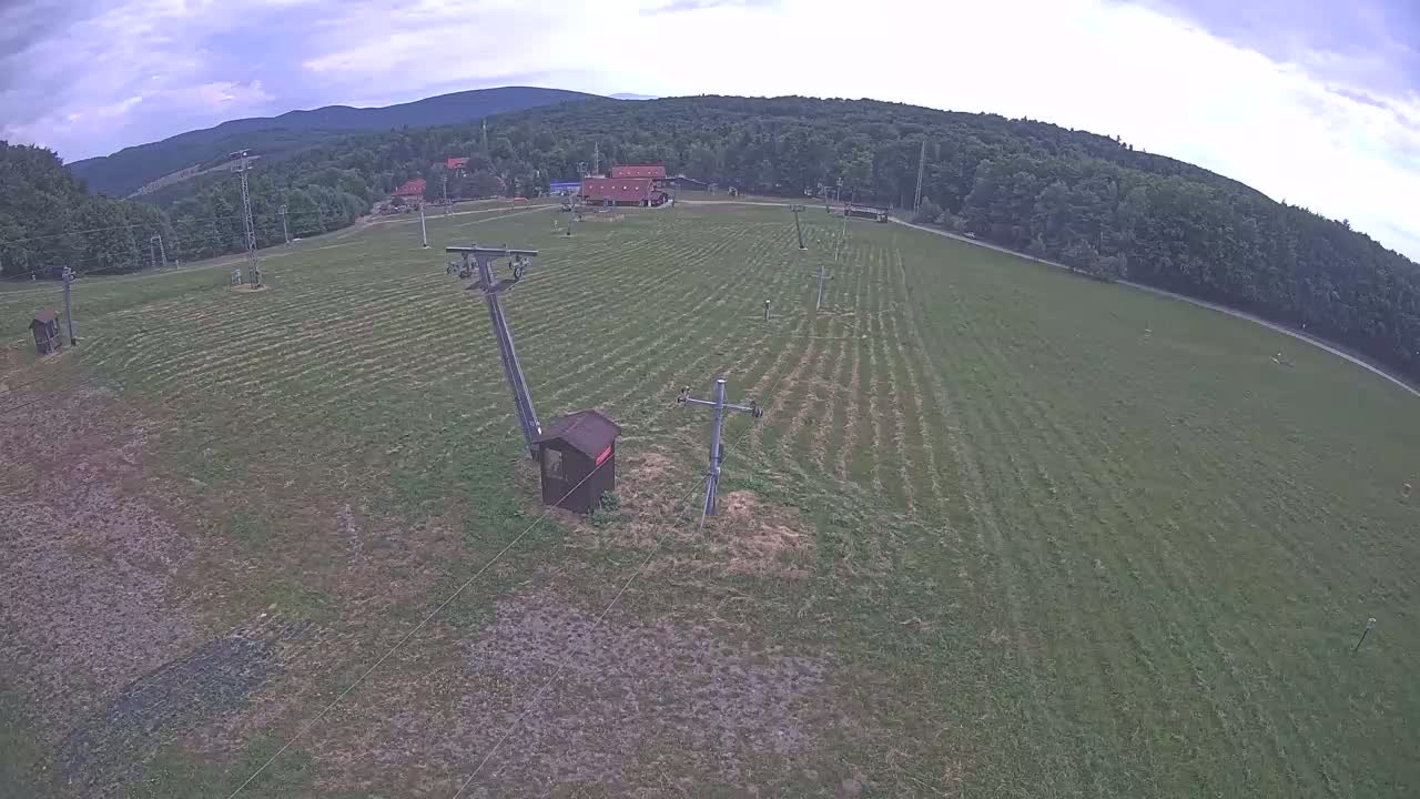 A grassy field with a ski lift infrastructure is visible under a partly cloudy sky, surrounded by a forest and distant mountains.