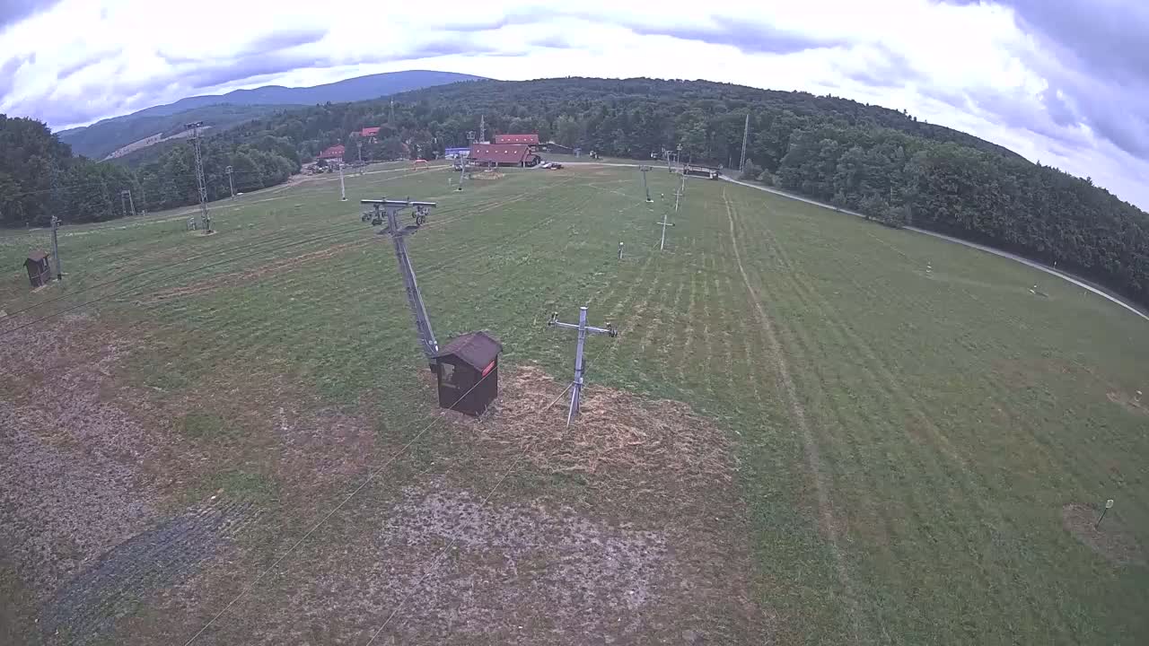 An aerial view shows a grassy field with ski lift infrastructure, a small building, and a line of trees and mountains in the background under a cloudy sky.