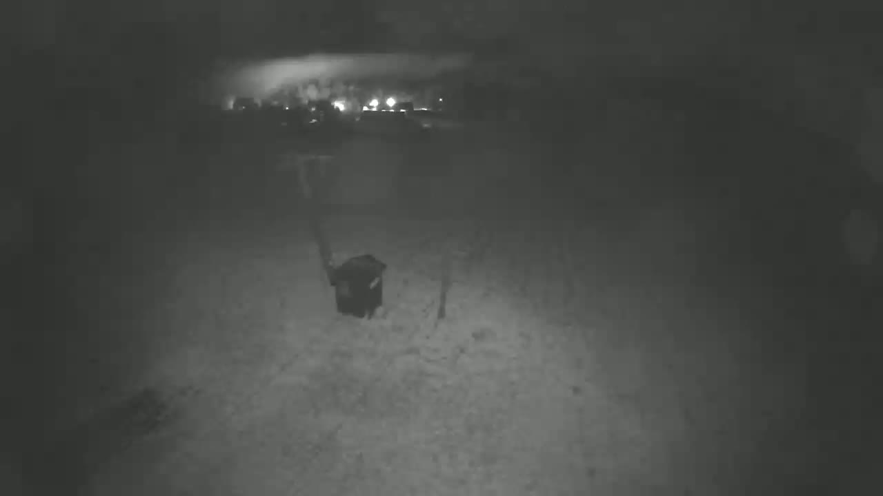 A dark, snowy nighttime scene shows a dark object, possibly a container, in the foreground with faint lights in the background.