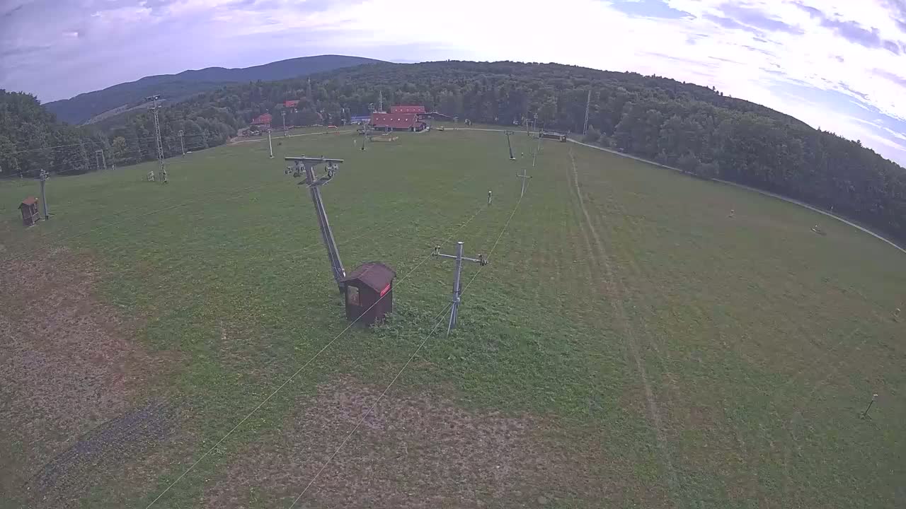 An aerial view shows a grassy field with a chairlift and a small building, under partly cloudy skies, backed by a line of trees and distant mountains.