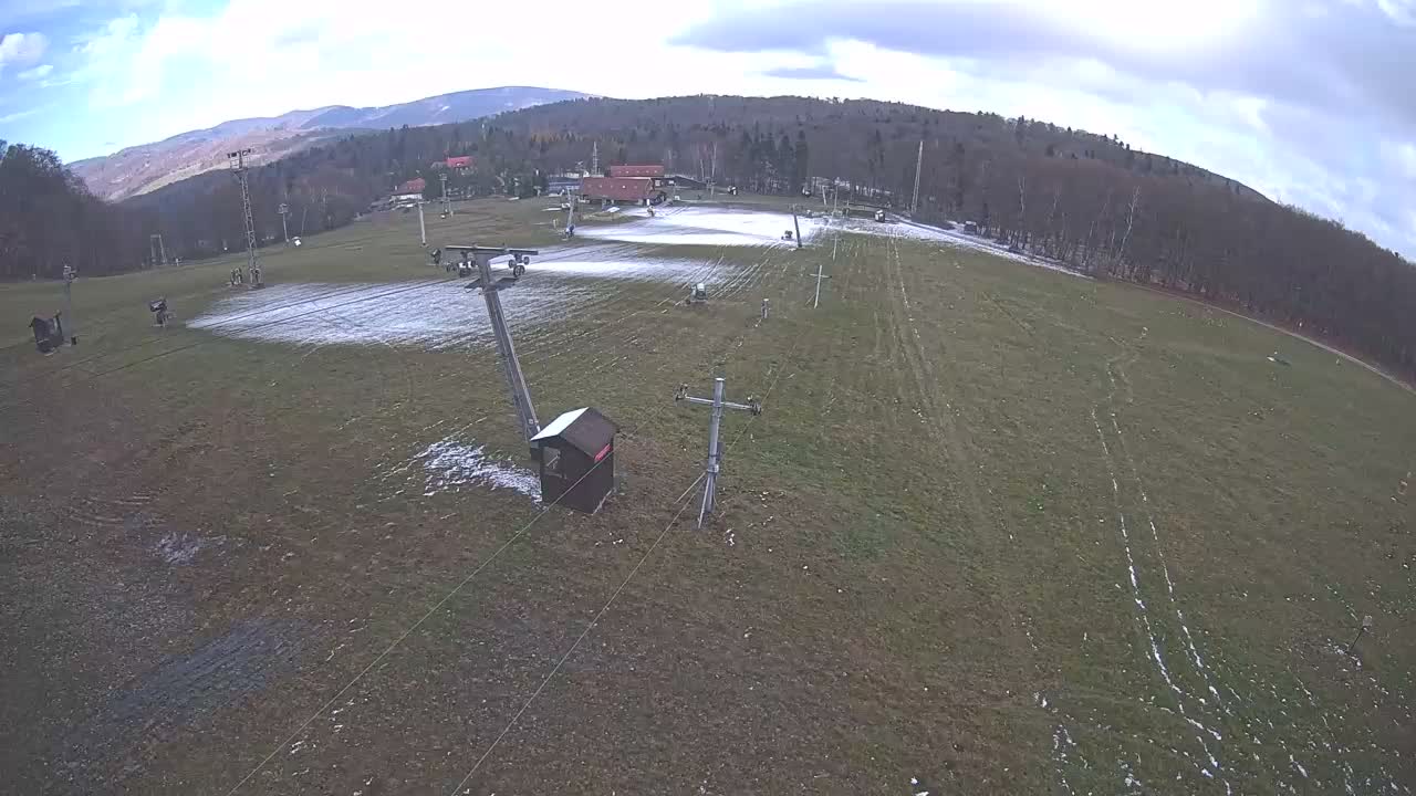 An aerial view shows a grassy field with a chairlift and a small building, under partly cloudy skies, backed by a line of trees and distant mountains.