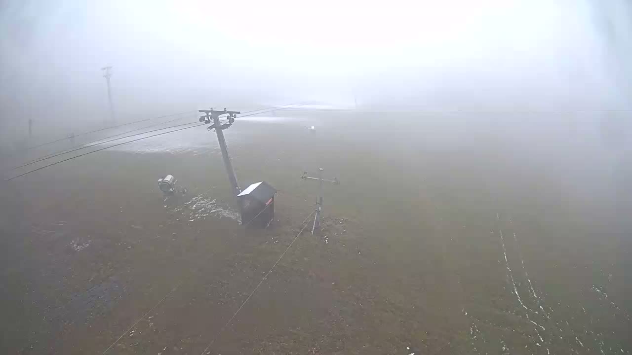A ski lift tower with cables, a snow-making machine actively spraying, a small building, and a weather station are visible on a muddy slope under very foggy conditions, with distant visibility heavily obscured.
