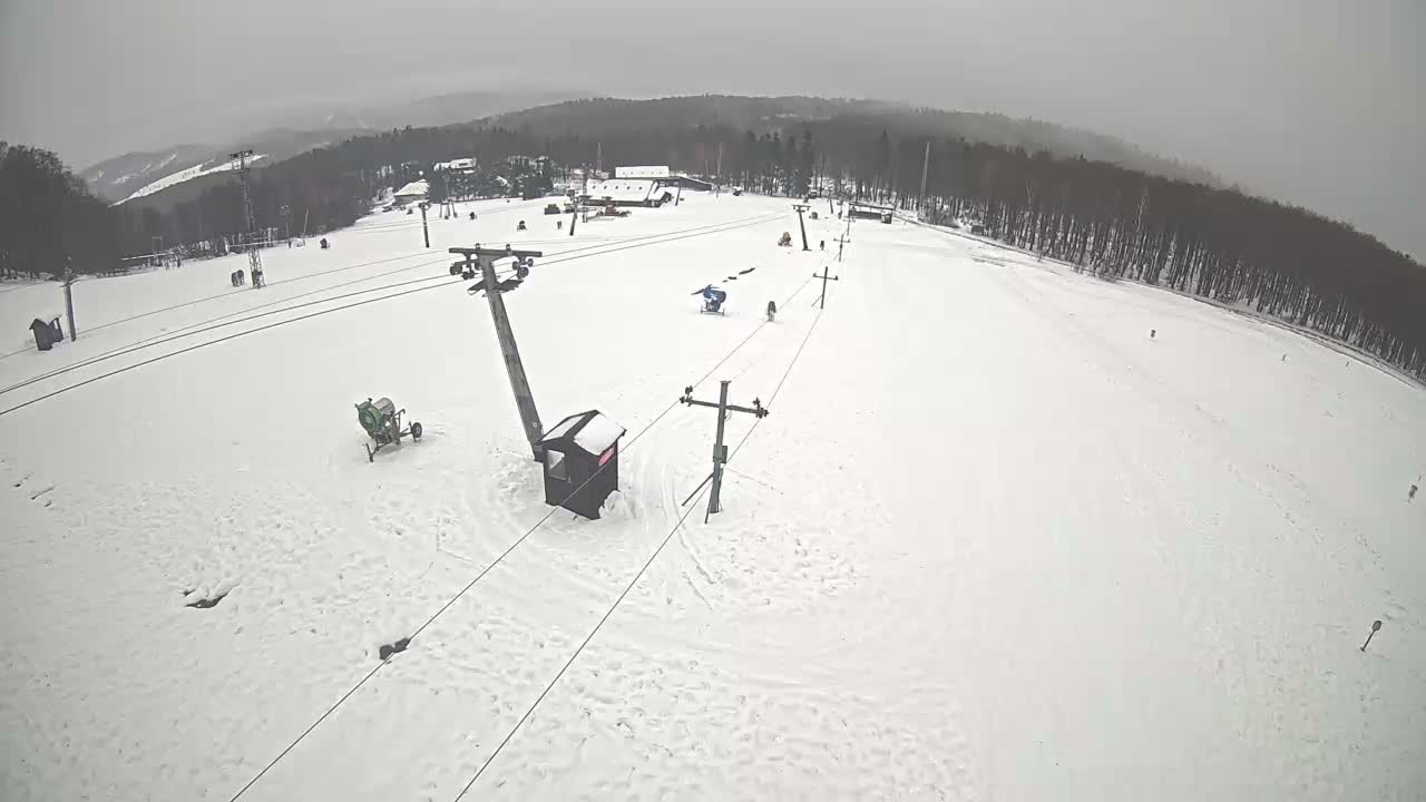 An overcast winter day reveals a snowy ski slope dotted with ski lift poles, a snow cannon, several distant buildings, and a few individuals skiing or snowboarding.