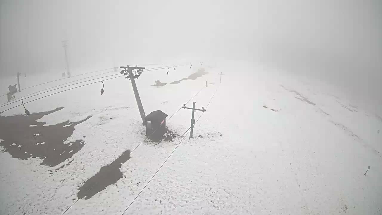 A snow-covered mountain landscape featuring ski lift poles and cables, with some patches of exposed ground, is obscured by dense fog and overcast skies.