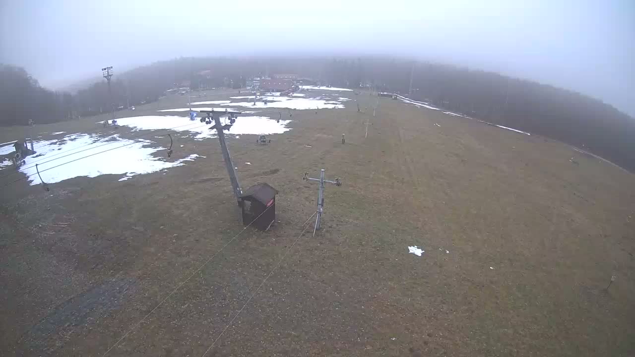 A snow-covered mountain landscape featuring ski lift poles and cables, with some patches of exposed ground, is obscured by dense fog and overcast skies.