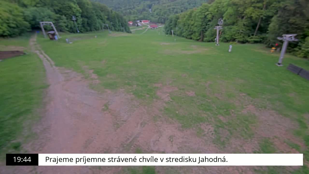 A grassy ski slope, mostly bare of snow, is visible with dirt paths and remnants of ski lifts against a backdrop of lush green trees.
