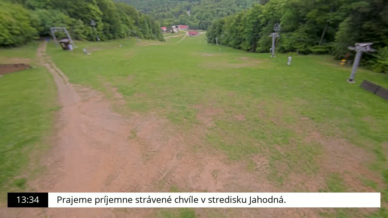 A grassy ski slope with a dirt path and chairlift infrastructure is visible, surrounded by a lush green forest under a seemingly clear sky.
