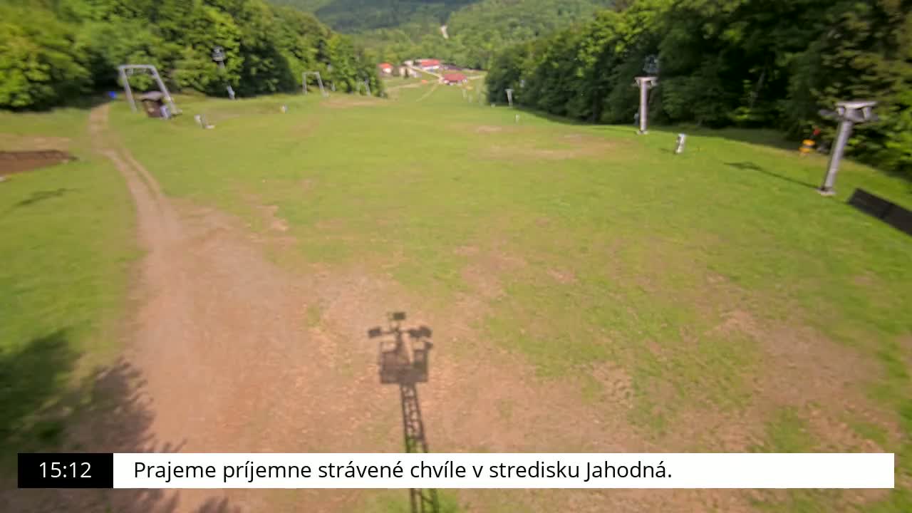 A grassy ski slope, mostly bare of snow, is visible from above on a sunny day, with a dirt path running alongside it and chairlift poles spaced along the sides.