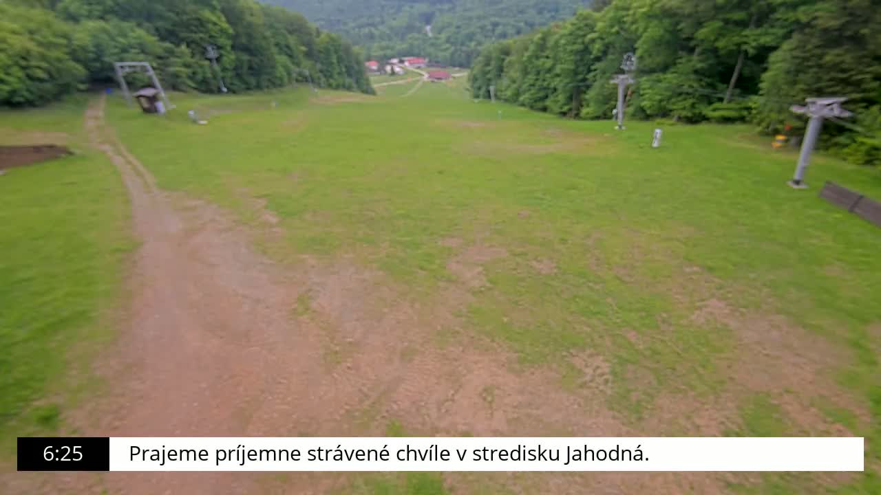 A grassy ski slope with two ski lifts and a dirt path leading to a small village in the distance, on a clear day.