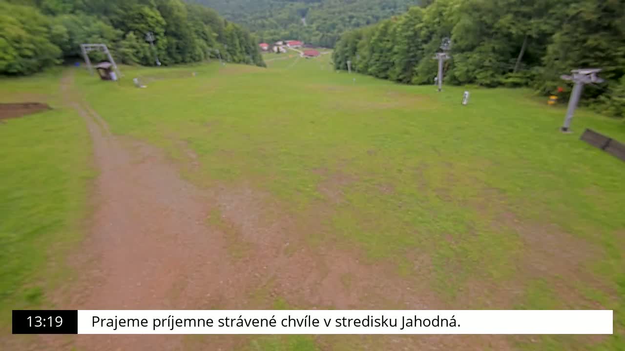 A grassy ski slope, possibly in the summer, with a dirt path leading to ski lifts and a small cluster of buildings in the distance.
