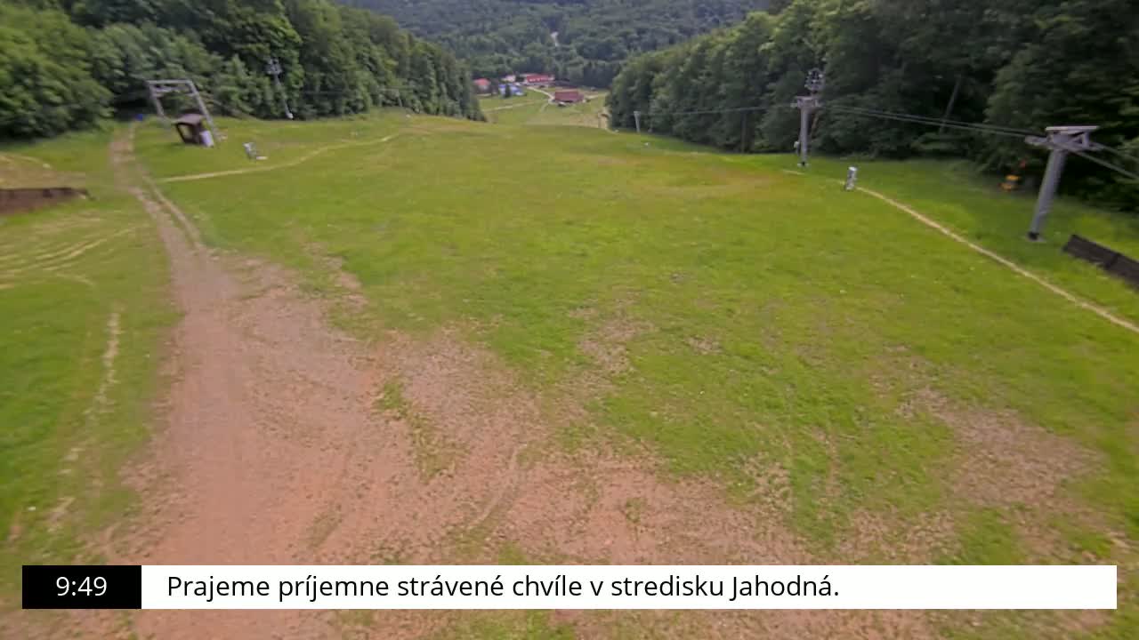 A grassy hill with a dirt path and ski lift infrastructure is visible, overlooking a small village nestled in a forest under a clear sky.