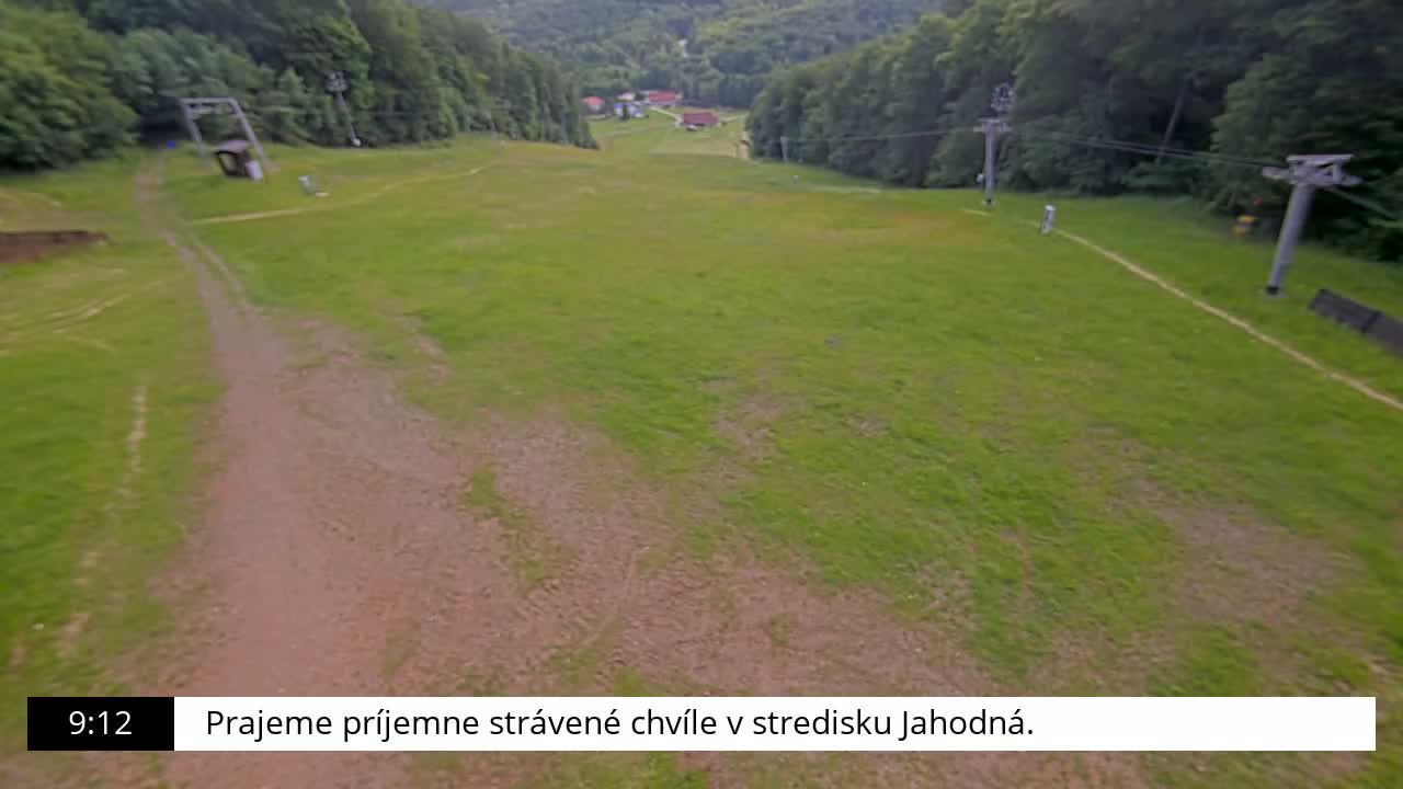 A grassy ski slope with a dirt path and chairlift towers is visible, surrounded by a lush green forest under a clear sky.