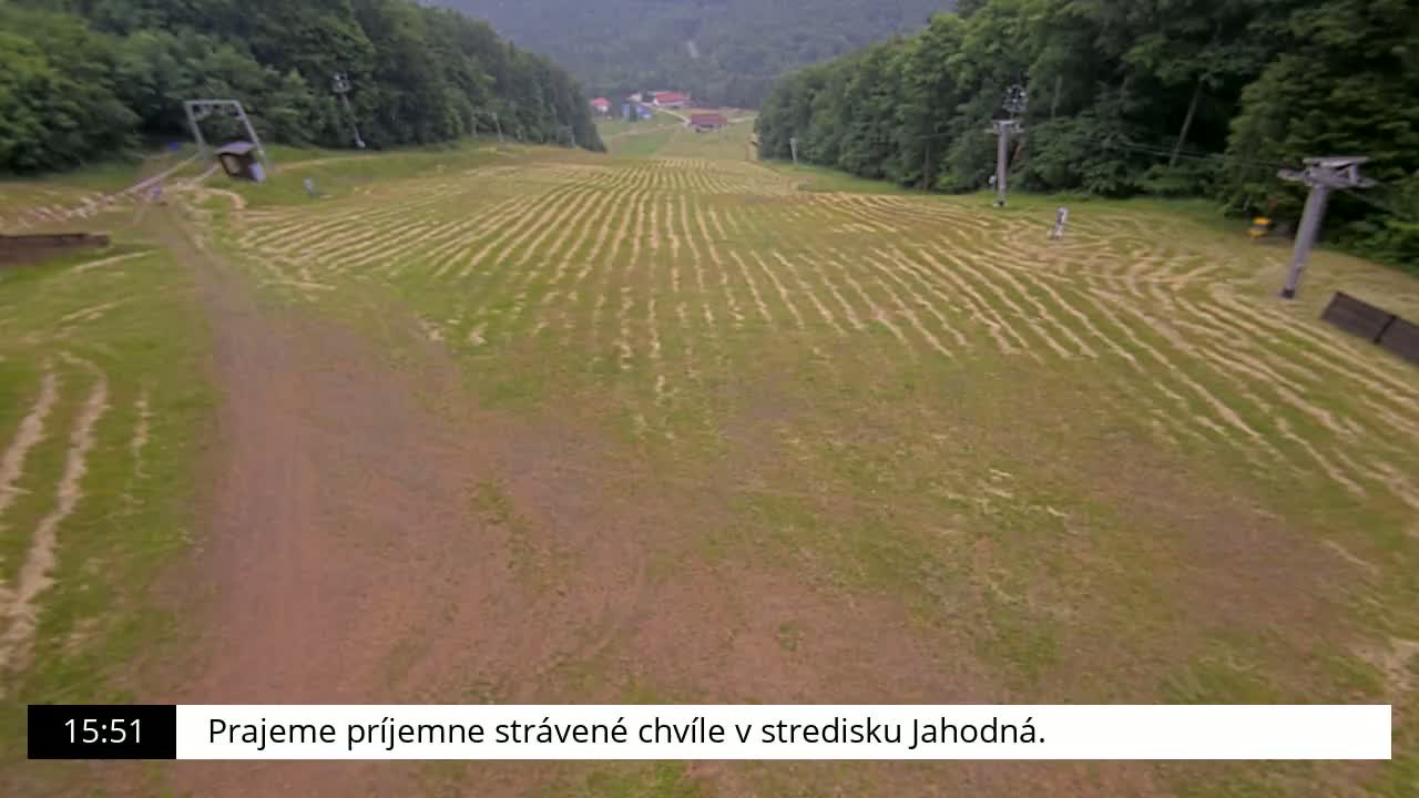 An expansive grassy field with parallel lines etched into the ground, surrounded by a forest and featuring two ski lifts, is viewed from above on an overcast day.