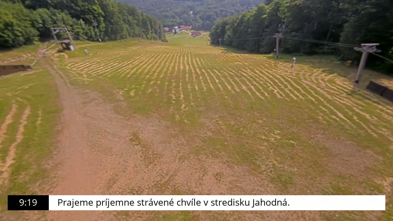 An aerial view shows a grassy hill with parallel lines cut into the grass, a dirt path, and a ski lift in the background on a sunny day.