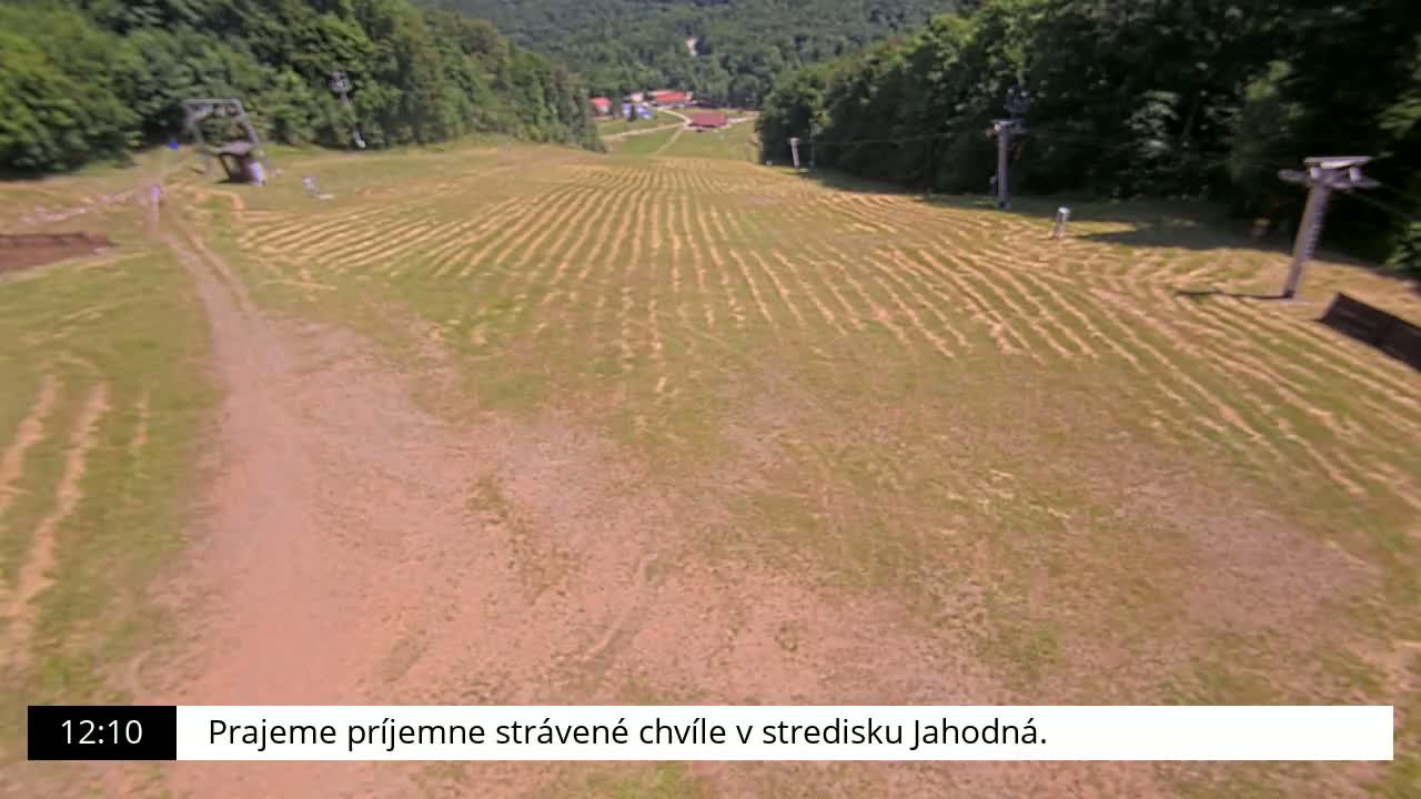A high-angle view shows a grassy hill with parallel lines cut into the grass, two ski lifts, and a small village in the background on a sunny day.