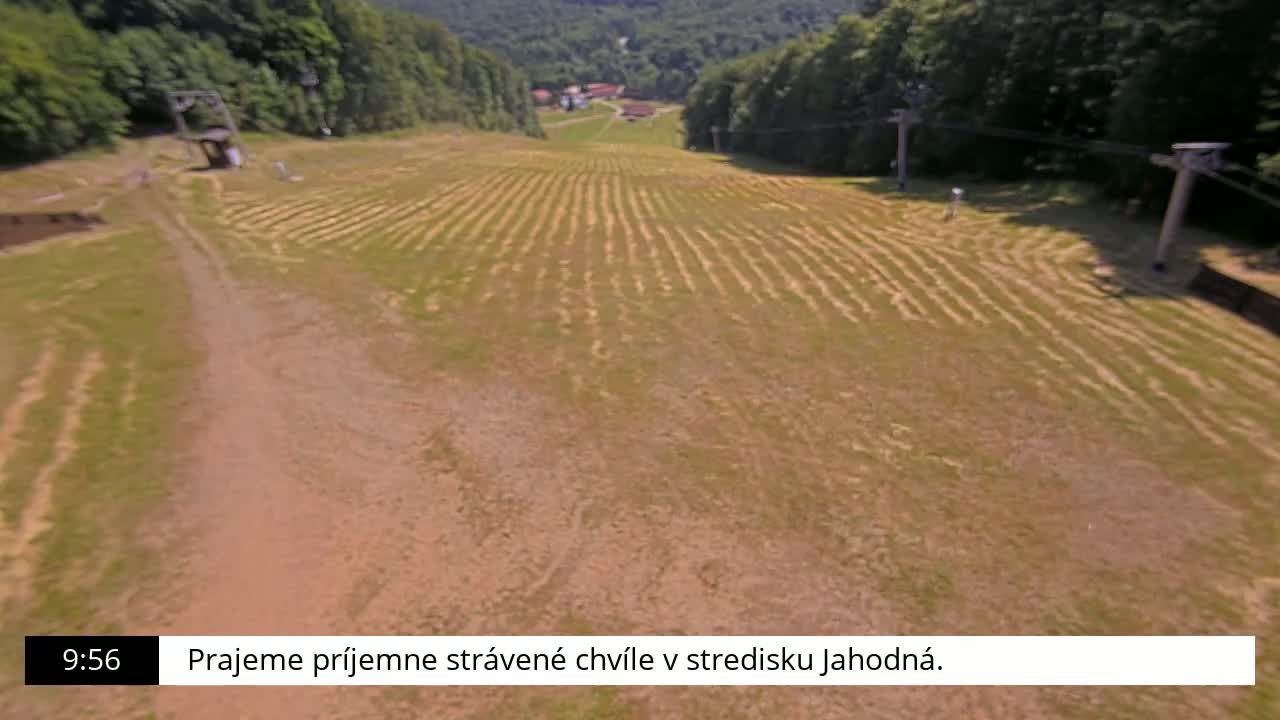 An aerial view shows a grassy, mostly barren ski slope with two chairlift stations and a dirt road leading up to them, under a sunny sky.