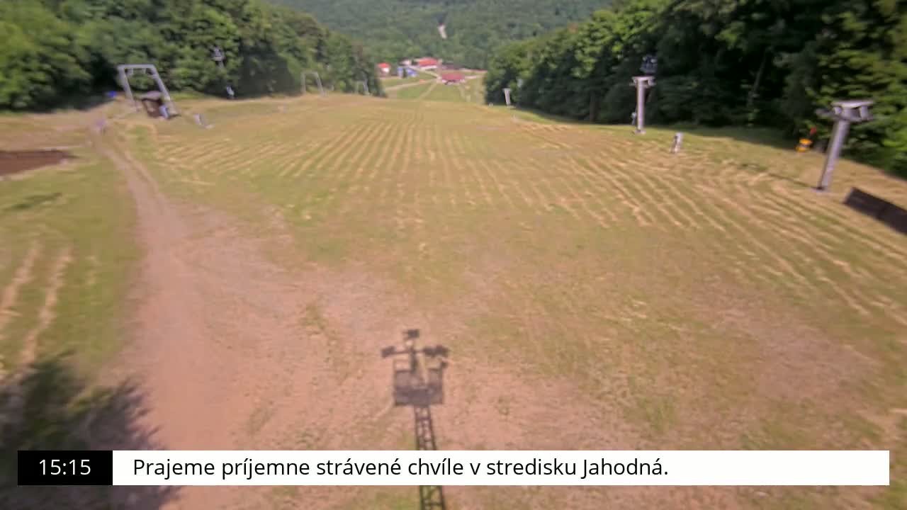 An aerial view shows a grassy ski slope with two chairlift towers on either side, surrounded by a green forest on a sunny day.