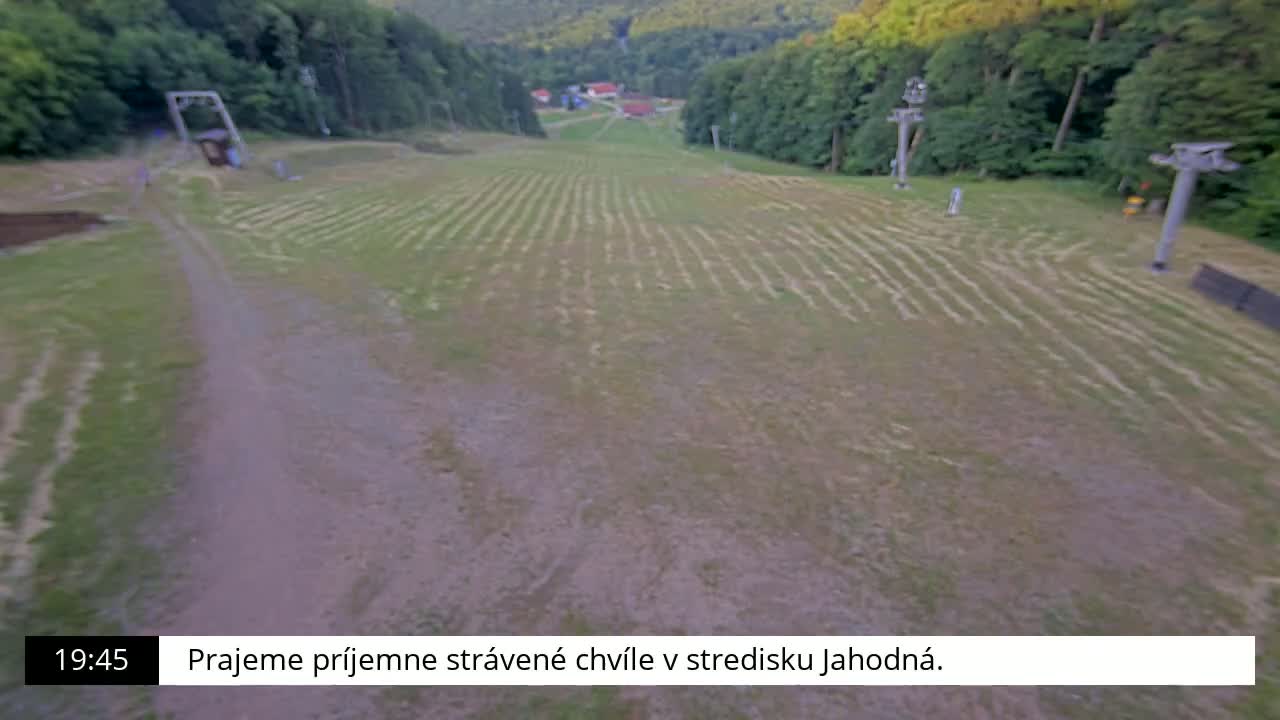A grassy, mostly barren ski slope is visible with some remnants of tire tracks or other markings, surrounded by a forest under a mostly clear sky.