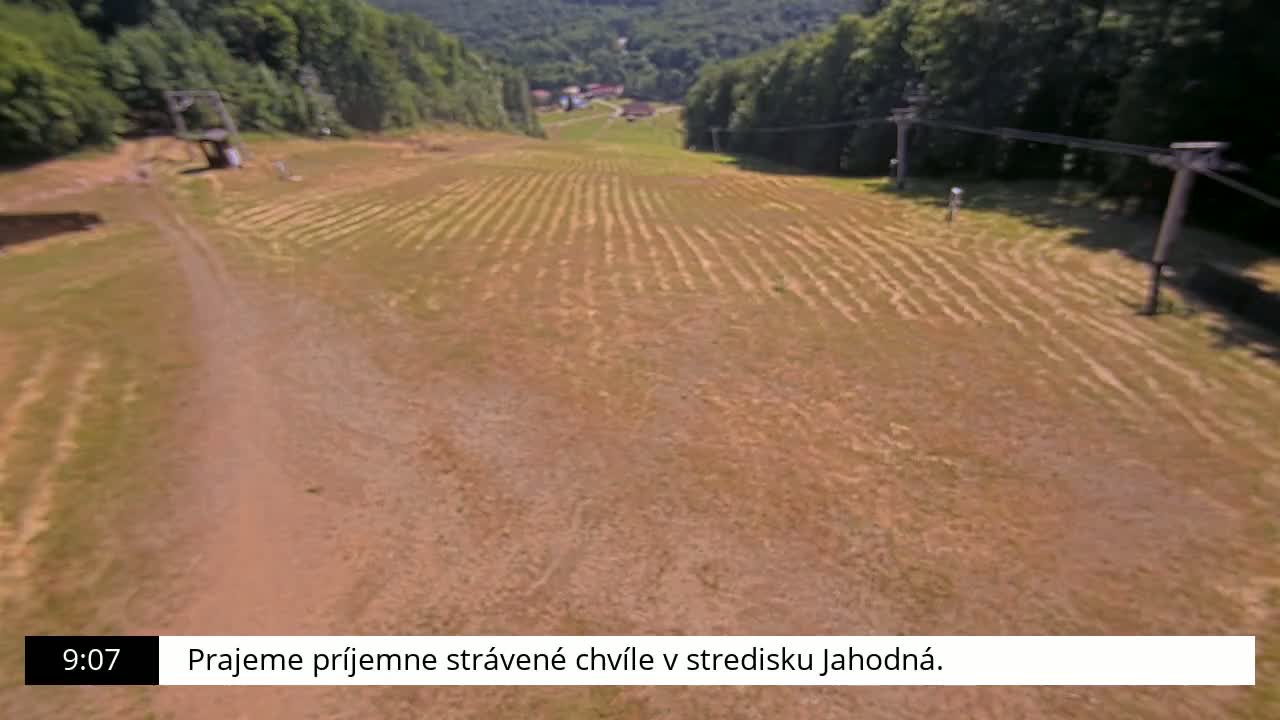 A grassy hill with parallel lines cut into it, possibly a ski slope in summer, is viewed from above on a sunny day.