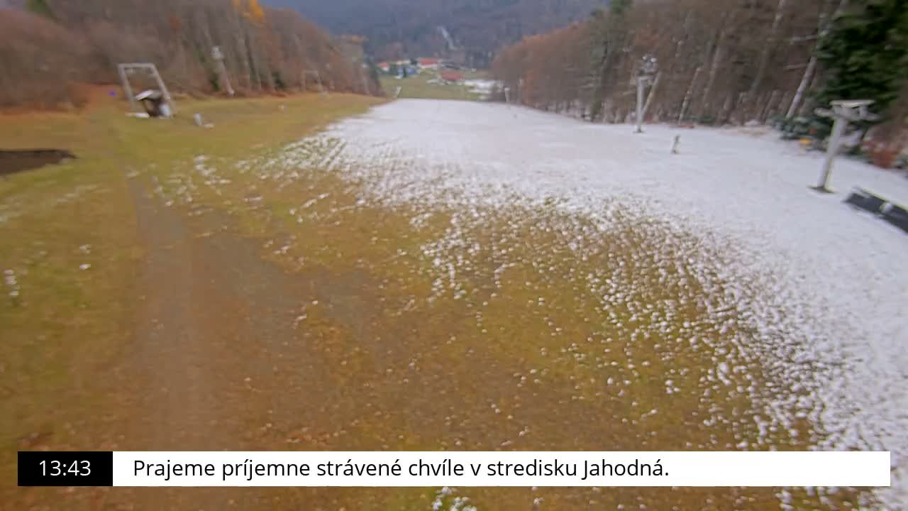 A person walks down a dirt path across a grassy hillside with ski lift infrastructure visible and a forested background on a cloudy day.