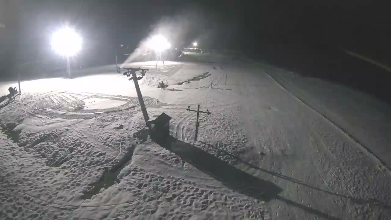 A brightly lit ski slope is seen at night under clear skies, with snow cannons actively making snow and tracks visible in the groomed and ungroomed snow.