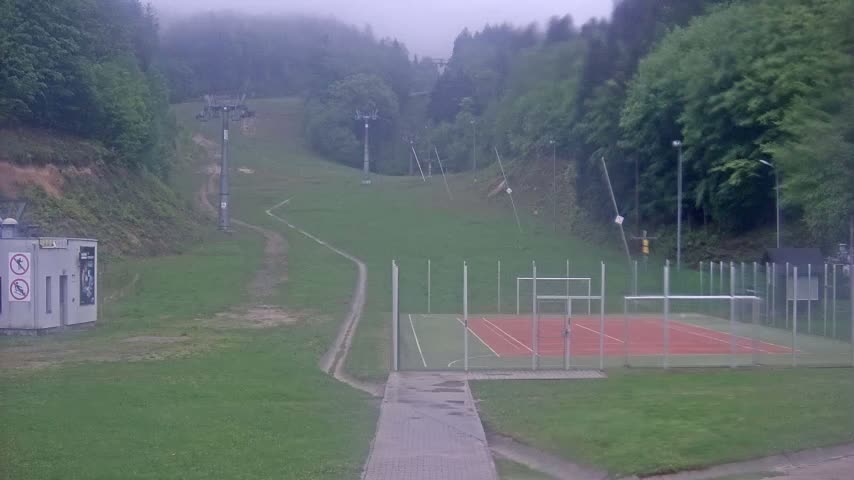 A foggy view of a grassy hillside with a ski lift, a small building, and an enclosed sports court.