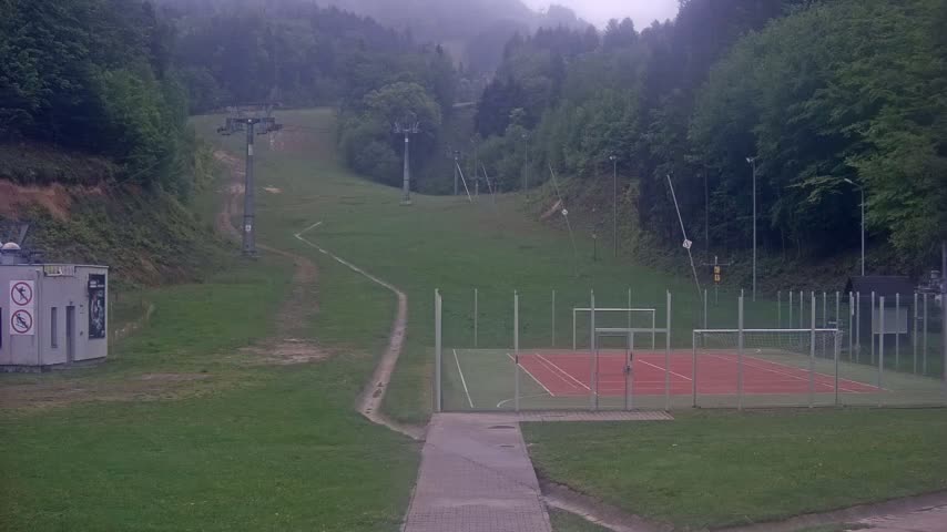 A paved walkway leads to an enclosed sports court situated on a grassy hillside, with a ski lift and forested mountains visible in the background on an overcast day.