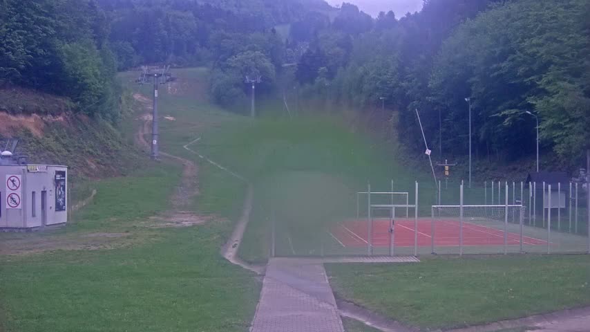 A hazy view shows a paved pathway leading to an enclosed tennis or handball court, with a ski lift and grassy hillside in the background.