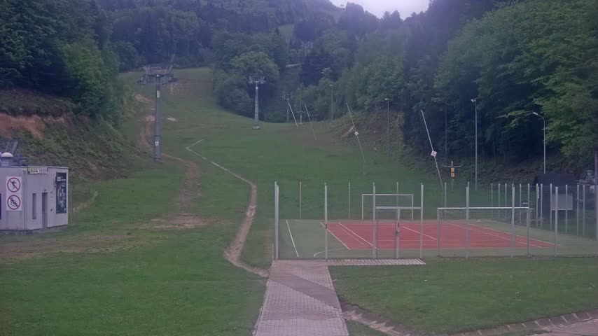A paved pathway leads to an outdoor sports court enclosed by a fence, situated below a grassy hillside with ski lift infrastructure and a forested background under an overcast sky.