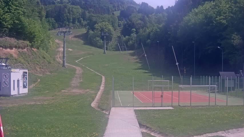 A paved walkway leads to a multi-purpose sports court enclosed by a fence, situated on a grassy hillside with a ski lift visible in the background on a sunny day.