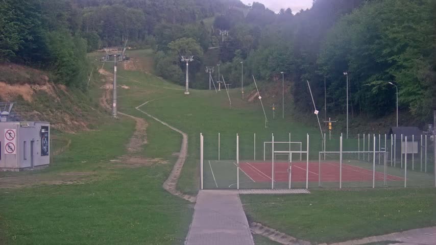 A paved walkway leads to a fenced multi-purpose court next to a grassy hillside with inactive ski lift infrastructure, under overcast skies.