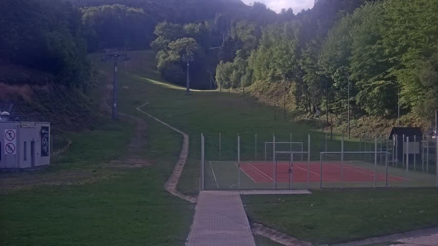 A paved walkway leads to an outdoor tennis court, situated on a grassy hillside below a ski lift and a wooded area on a partly sunny day.