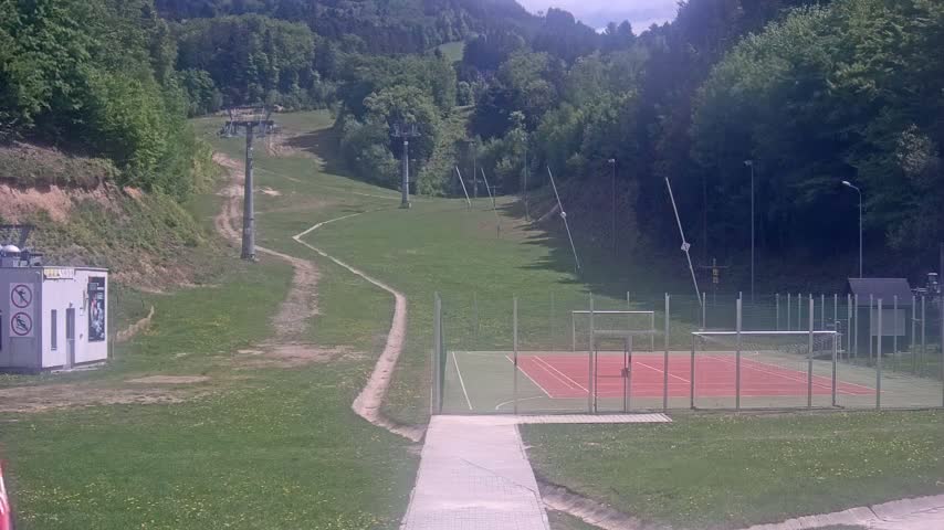 A paved pathway leads to a red and green sports court enclosed by a fence, situated on a grassy hillside below a ski lift and a forested mountain under a partly cloudy sky.