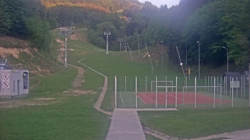 A grassy hillside with ski lifts and a small, fenced-in sports court is visible under an overcast sky.