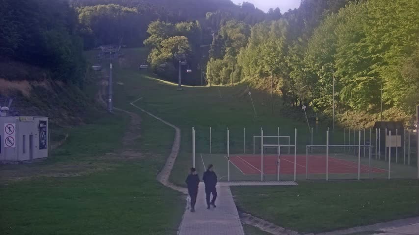 Two people walk a small dog on a paved path near an outdoor tennis court and a grassy hillside with a chairlift in the background under a partly sunny sky.