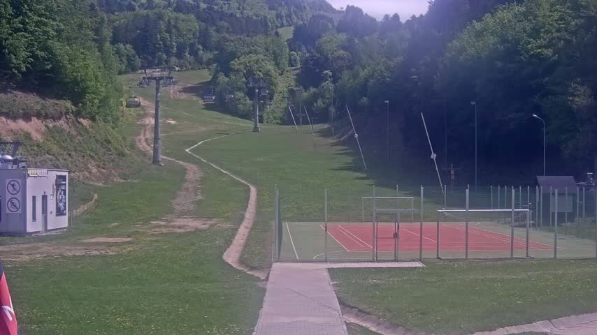 A grassy hillside with a chairlift, a paved walkway leading to a multi-use sports court, all under a partly sunny sky.