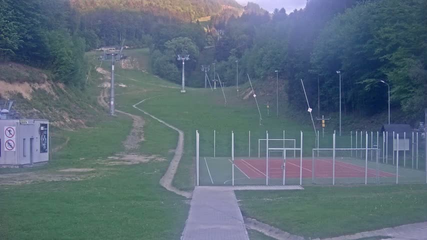 A grassy hillside with ski lift infrastructure and an enclosed sports court is visible, under an overcast sky.