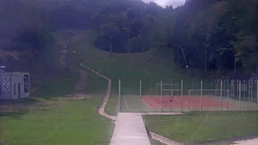 A paved pathway leads to an outdoor tennis and basketball court surrounded by a chain-link fence, situated below a grassy hillside with a ski lift and forested background under an overcast sky.