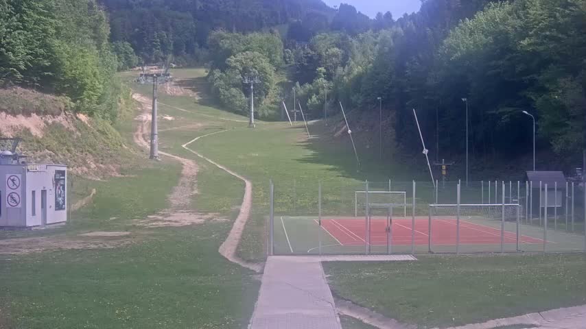 A grassy hillside with a ski lift, a paved pathway leading to an enclosed multi-use games area, and a small building are visible on a partly sunny day.