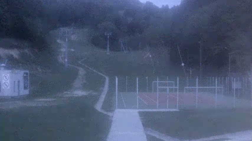 A hazy view shows a paved path leading to an outdoor basketball court enclosed by a fence, with a ski lift and surrounding hills visible in the background.