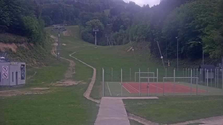 A grassy hillside with ski lift infrastructure, a small building, and an enclosed multi-purpose sports court is visible under an overcast sky.