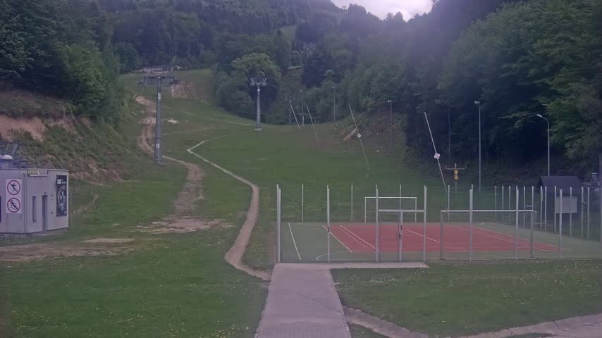 A grassy hillside with ski lifts, a dirt path, and a fenced-in sports court is visible on an overcast day.