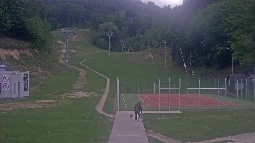 A person walks a dog on a paved path toward a small, red outdoor court surrounded by a fence, with a grassy hillside featuring ski lifts and trees in the background, under overcast skies.