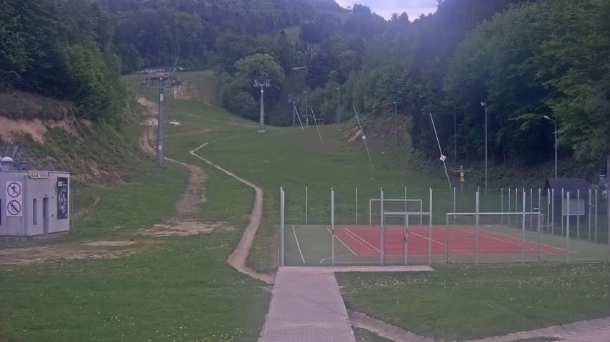A multi-use games area sits at the base of a grassy hill with ski lifts and a winding trail.