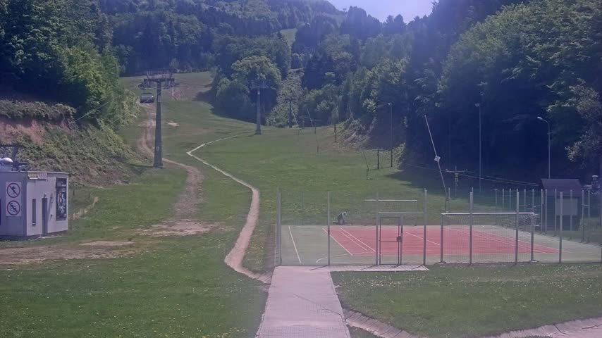 A paved pathway leads to an outdoor tennis court situated on a grassy area below a ski lift on a sunny day amidst a hilly, wooded landscape.